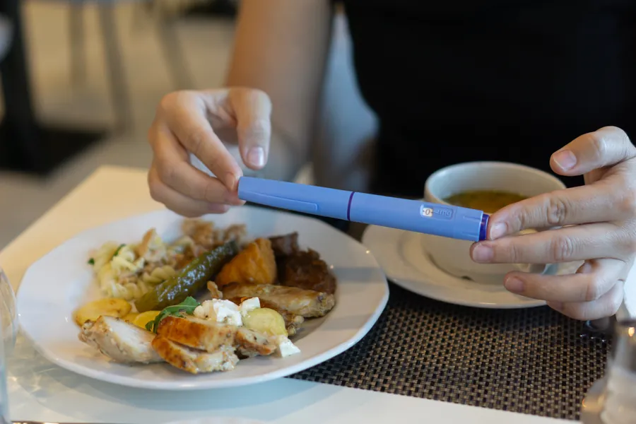 Close-up of hands holding an Ozempic injection pen while sitting at a table with a plate of food and a cup of soup.